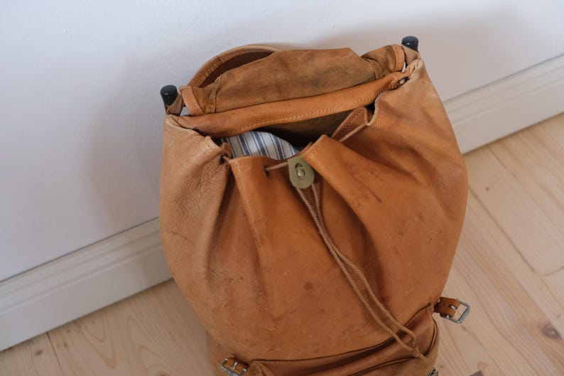 May include: A vintage, open leather backpack in a warm tan color. The bag features a drawstring closure with a brass clasp and adjustable straps. The interior reveals a glimpse of striped fabric. The backpack is designed with a classic, rustic aesthetic.
