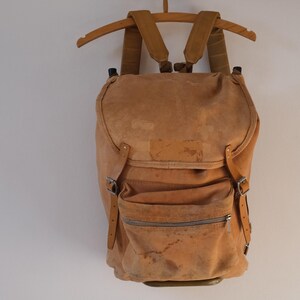 May include: A tan suede backpack with adjustable straps and a front zippered pocket. The backpack is hanging on a wooden hanger against a white wall. The bag has a vintage, worn appearance.