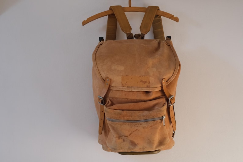 May include: A tan suede backpack with adjustable straps and a front zippered pocket. The backpack is hanging on a wooden hanger against a white wall. The bag has a vintage, worn appearance.