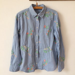 May include: Light blue button-down shirt with long sleeves and a single chest pocket. The shirt is adorned with embroidered floral patterns in shades of orange, green, and white. The shirt hangs on a wooden hanger against a white background.