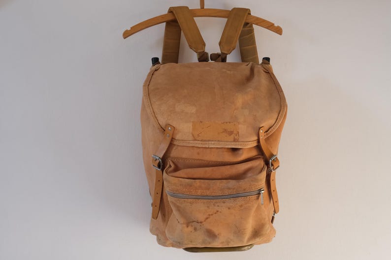 May include: A tan suede backpack with adjustable straps and a front zippered pocket. The vintage bag is suspended from a wooden hanger against a white background. The backpack has a flap closure and metal buckles.