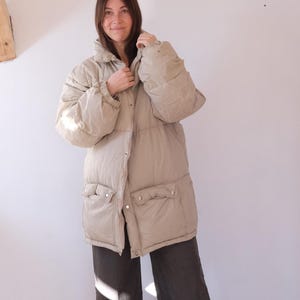 Könnte beinhalten: Eine beige Steppjacke mit hohem Kragen und zwei Vordertaschen. Die Jacke hat einen Knopfverschluss und lange Ärmel. Das Model trägt eine dunkelgraue Hose. Die Jacke scheint aus einem gesteppten Material zu sein.