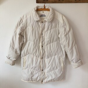 May include: A white quilted puffer jacket featuring a fold-down collar, long sleeves with cuffs, and a front closure with silver-toned metal snap buttons. The jacket has a chevron-patterned stitching design and two side pockets on the front.
