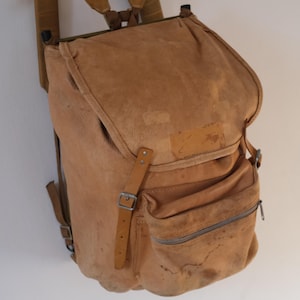 May include: A vintage-style backpack in a light brown hue, featuring a flap closure and a front zippered pocket. The bag has adjustable straps and a buckle closure. The backpack is made of a worn, textured material, and is hanging on a wooden hanger.