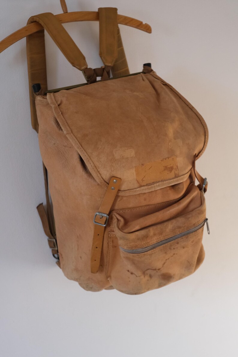 May include: A vintage-style backpack in a light brown hue, featuring a flap closure and a front zippered pocket. The bag has adjustable straps and a buckle closure. The backpack is made of a worn, textured material, and is hanging on a wooden hanger.