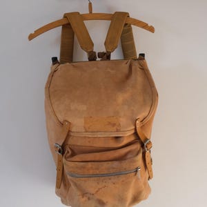 May include: A tan-colored backpack with adjustable straps and a front zippered pocket. The vintage-style bag is made of a worn, textured material and hangs from a wooden hanger. The backpack has a flap closure and metal buckles.