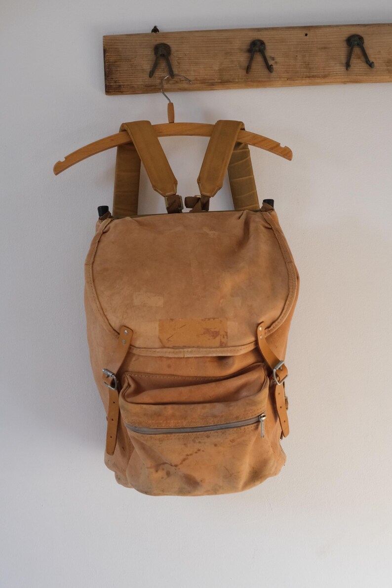 May include: A tan-colored backpack with adjustable straps and a front zippered pocket. The vintage-style bag is made of a worn, textured material and hangs from a wooden hanger. The backpack has a flap closure and metal buckles.