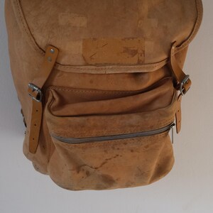 May include: A worn, tan leather backpack with a flap closure and a front zippered pocket. The bag features adjustable straps with silver buckles and a distressed appearance, suggesting age and use. The bag is designed for carrying items.