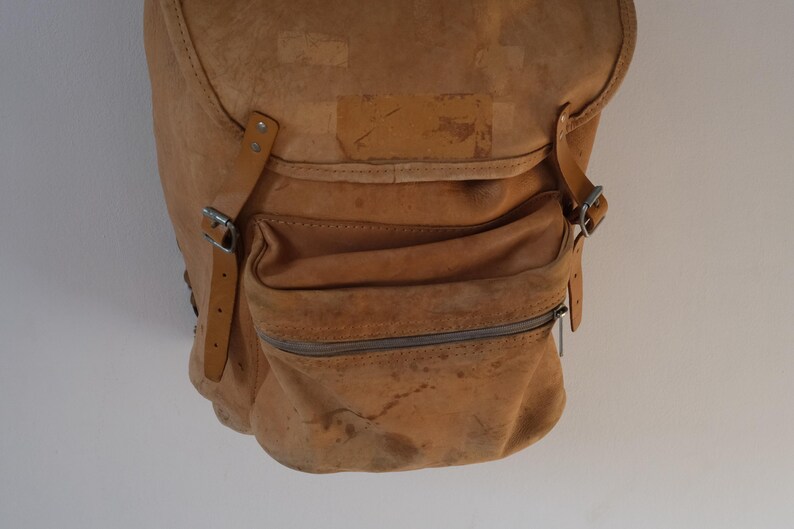 May include: A worn, tan leather backpack with a flap closure and a front zippered pocket. The bag features adjustable straps with silver buckles and a distressed appearance, suggesting age and use. The bag is designed for carrying items.