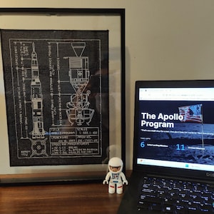 K&ouml;nnte beinhalten: Eine gerahmte Schwarzwei&szlig;illustration der Apollo-Programm Saturn V Rakete und des Mondmoduls. Die Illustration ist auf schwarzem Hintergrund und enth&auml;lt technische Details. Ein Laptop-Computer ist auf einer Website &uuml;ber das Apollo-Programm ge&ouml;ffnet. Die Website zeigt ein Bild eines Astronauten auf dem Mond und den Text "The Apollo Program". Eine kleine Astronautenfigur befindet sich im Vordergrund.