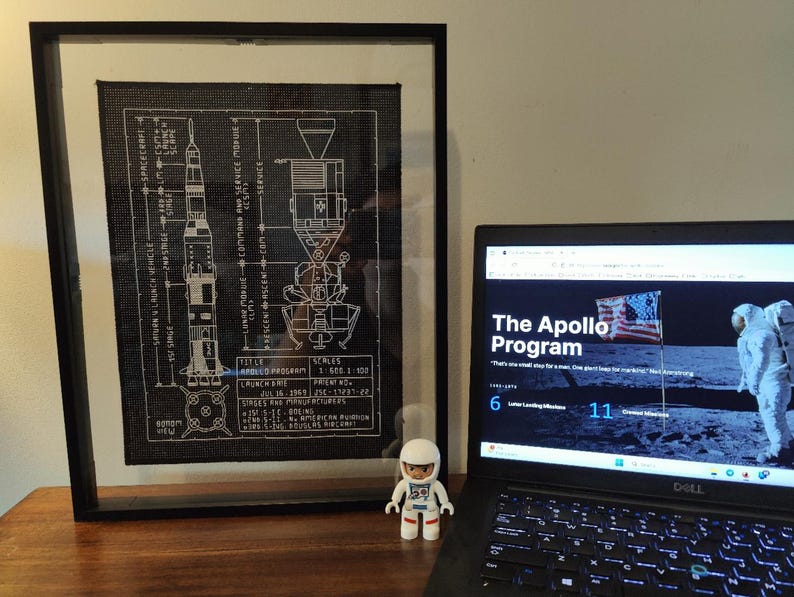 K&ouml;nnte beinhalten: Eine gerahmte Schwarzwei&szlig;illustration der Apollo-Programm Saturn V Rakete und des Mondmoduls. Die Illustration ist auf schwarzem Hintergrund und enth&auml;lt technische Details. Ein Laptop-Computer ist auf einer Website &uuml;ber das Apollo-Programm ge&ouml;ffnet. Die Website zeigt ein Bild eines Astronauten auf dem Mond und den Text "The Apollo Program". Eine kleine Astronautenfigur befindet sich im Vordergrund.