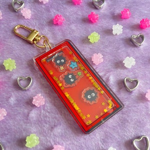 May include: A red and gold keychain with a clear plastic charm featuring three black and white cartoon characters with stars and a blue star. The keychain has a gold metal clasp.