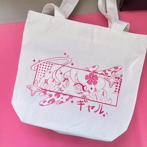 May include: White canvas tote bag featuring a pink anime-style illustration. The design includes a girl with a halo, stars, and Japanese text. The bag has two white handles and is set against a pink background.