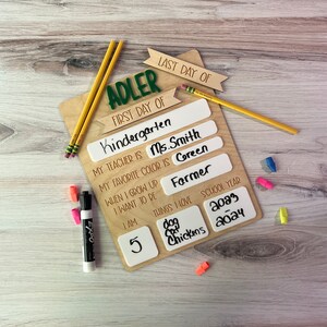 May include: A wooden sign with the name "Adler" and the words "First Day of Kindergarten" and "Last Day of" on it. The sign has spaces for the child to fill in information about their school year, including their teacher's name, favorite color, what they want to be when they grow up, and things they love. The sign also includes a space for the child's age and the school year.