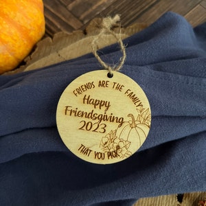 May include: A round wooden ornament with the text "Friends are the family Happy Friendsgiving 2023 That you pick" and a drawing of a pumpkin and flowers.