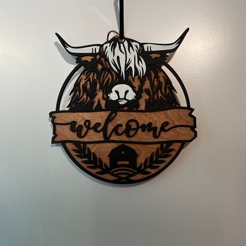 May include: A wooden welcome sign with a black silhouette of a Highland cow's head. The sign is round and has a brown wood grain finish. The words "welcome" are written in black cursive script on a brown banner.