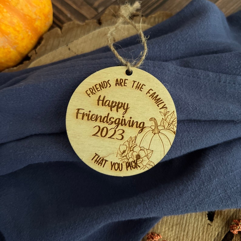 May include: A round wooden ornament with the text "Friends are the family Happy Friendsgiving 2023 That you pick" and a drawing of a pumpkin and flowers.