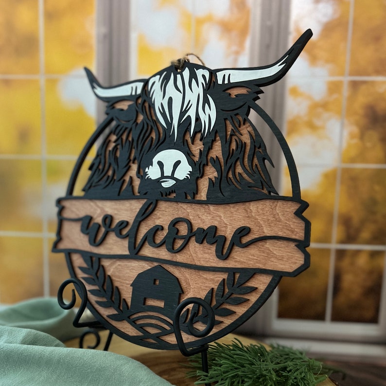 May include: Black wooden welcome sign with a cutout of a Highland cow head and the word "welcome" on a woodgrain background. The sign is on a black metal stand.