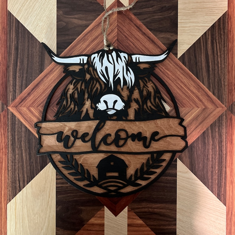 May include: A wooden welcome sign with a black silhouette of a Highland cow's head and the word "welcome" written in a cursive font. The sign is decorated with a small barn and a wreath of leaves.