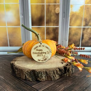 May include: A round wooden ornament with the text "Friends are the family. Happy Friendsgiving 2023 that you pick" and a small pumpkin design. The ornament is hanging from a string and is resting on a wooden surface.
