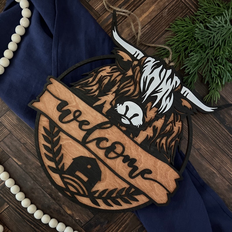 May include: Wooden welcome sign with a black silhouette of a Highland cow and the word "welcome" in a decorative script font. The sign has a brown wood grain background and a black barn silhouette in the lower left corner.