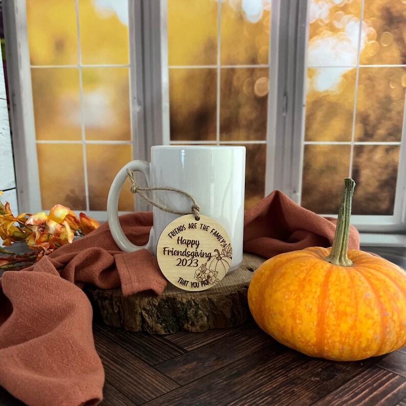 May include: A round wooden ornament with the text "Friends are the family you choose. Happy Friendsgiving 2023" and a pumpkin design. The ornament is hanging from a white mug on a wooden surface with a pumpkin and a brown fabric.