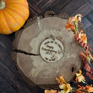 May include: A round wooden ornament with the text "Friends are the family Happy Friendsgiving 2023 That you pick" and a drawing of a pumpkin and flowers.