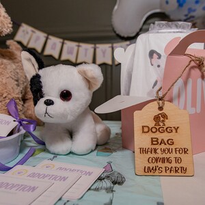 Peut inclure: Un chiot en peluche blanc et noir est assis pr&egrave;s d'un sac cadeau rose avec le nom "Olivia". Une &eacute;tiquette en bois indique "Doggy Bag Thank you for coming to Liv's Party". Des cartes d'adoption et un chien en peluche sont &eacute;galement visibles.