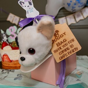 Peut inclure: Une t&ecirc;te de chien en peluche blanche et noire sort d'une bo&icirc;te rose, avec une &eacute;tiquette en bois indiquant "DOGGY BAG THANK YOU FOR COMING TO LIVY'S PARTY". Un ruban violet et un g&acirc;teau aux fraises sont &eacute;galement visibles.