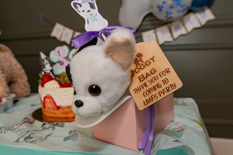 Peut inclure: Une t&ecirc;te de chien en peluche blanche et noire sort d'une bo&icirc;te rose, avec une &eacute;tiquette en bois indiquant "DOGGY BAG THANK YOU FOR COMING TO LIVY'S PARTY". Un ruban violet et un g&acirc;teau aux fraises sont &eacute;galement visibles.
