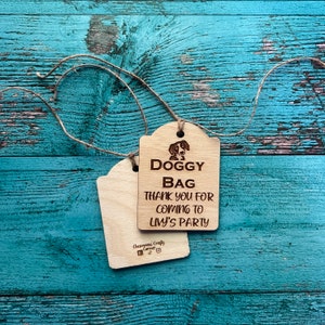 Peut inclure: Deux &eacute;tiquettes en bois pour sacs &agrave; chien avec de la ficelle. Les &eacute;tiquettes ont une finition en bois clair et pr&eacute;sentent une illustration de chien et le texte "DOGGY BAG THANK YOU FOR COMING TO LIVY'S PARTY". Les &eacute;tiquettes sont sur une surface en bois bleu-vert vieilli.