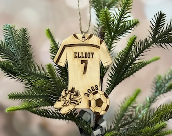 May include: A wooden soccer jersey ornament with the name "Elliot" and the number "7" on the back. The ornament also features a soccer ball and cleats, and the year "2022" is engraved on the bottom.