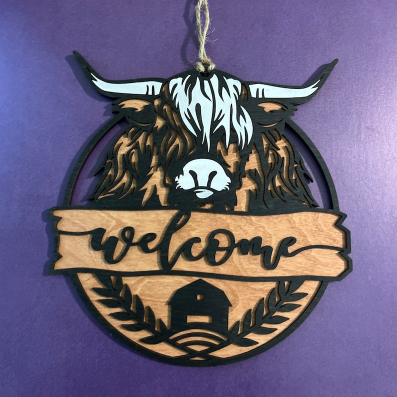 May include: A wooden welcome sign featuring a black silhouette of a Highland cow with white markings. The sign has a brown woodgrain background with the word "welcome" in black script. A small black silhouette of a barn is at the bottom of the sign.