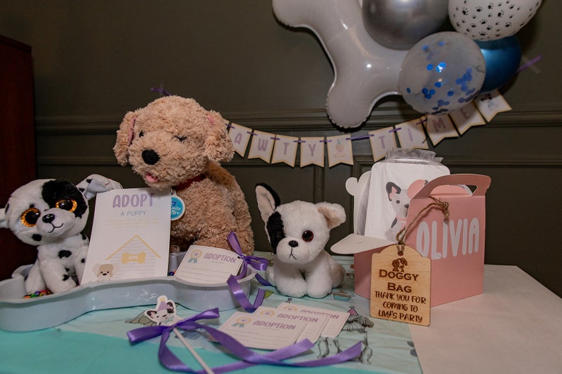 Peut inclure: Une sc&egrave;ne de f&ecirc;te avec des peluches de chiots, des cadeaux de f&ecirc;te et des ballons. La sc&egrave;ne comprend une bo&icirc;te cadeau rose avec le nom "Olivia", une &eacute;tiquette en bois avec "Doggy Bag" et des panneaux "Adoptez un chiot".