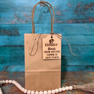 Peut inclure: Un sac cadeau en papier brun avec une &eacute;tiquette indiquant "Doggy Bag Thank you for coming to Livy's Party". L'&eacute;tiquette pr&eacute;sente une illustration de chien. Le sac a une poign&eacute;e en papier torsad&eacute;.