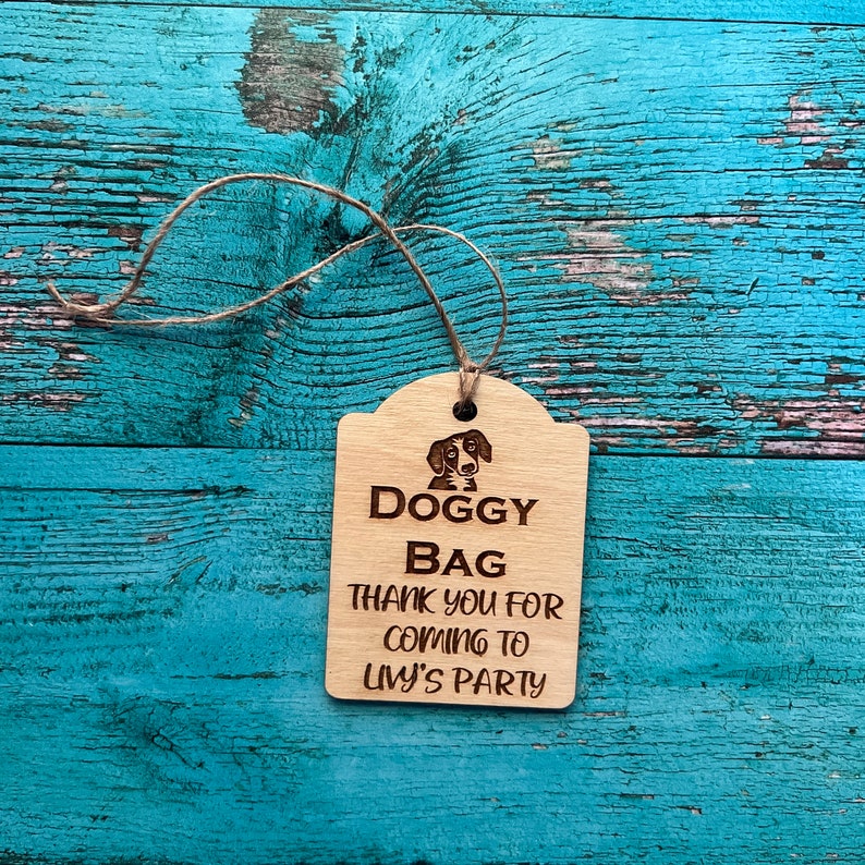 Peut inclure: Une &eacute;tiquette en bois "Doggy Bag" avec une ficelle. L'&eacute;tiquette pr&eacute;sente une illustration de chien et le texte "Thank you for coming to Livy's Party". L'&eacute;tiquette est en bois clair et est pos&eacute;e sur un fond turquoise.