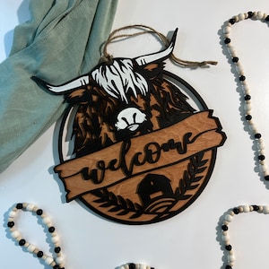 May include: A wooden welcome sign with a black silhouette of a highland cow and the word "welcome" in a brown banner. The sign is decorated with a small house and greenery.