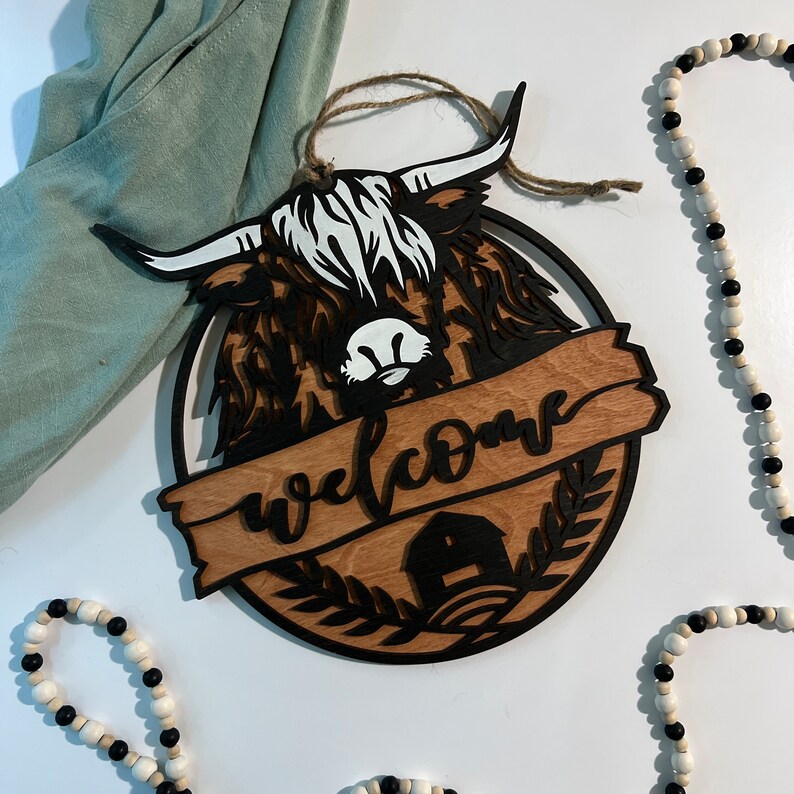 May include: A wooden welcome sign with a black silhouette of a highland cow and the word "welcome" in a brown banner. The sign is decorated with a small house and greenery.
