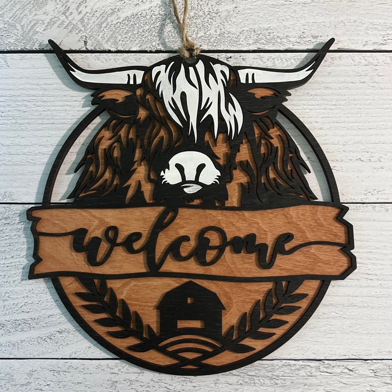 May include: A wooden welcome sign featuring a black silhouette of a Highland cow with a white face and a brown and tan wooden background. The sign has the word "welcome" written in black cursive lettering and a small black barn silhouette at the bottom.
