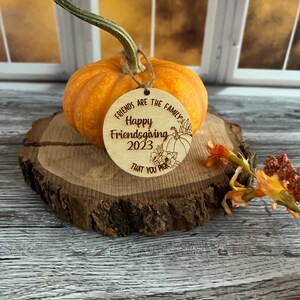 May include: A round wooden ornament with the text "Friends are the family Happy Friendsgiving 2023 that you pick" and a small pumpkin illustration.