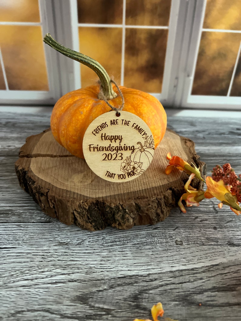 May include: A round wooden ornament with the text "Friends are the family Happy Friendsgiving 2023 that you pick" and a small pumpkin illustration.