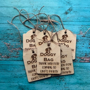 Peut inclure: Plusieurs &eacute;tiquettes cadeaux en bois avec une illustration de chien et les mots "DOGGY BAG" et "THANK YOU FOR COMING TO LIVY'S PARTY" grav&eacute;s. Chaque &eacute;tiquette est attach&eacute;e avec de la ficelle.