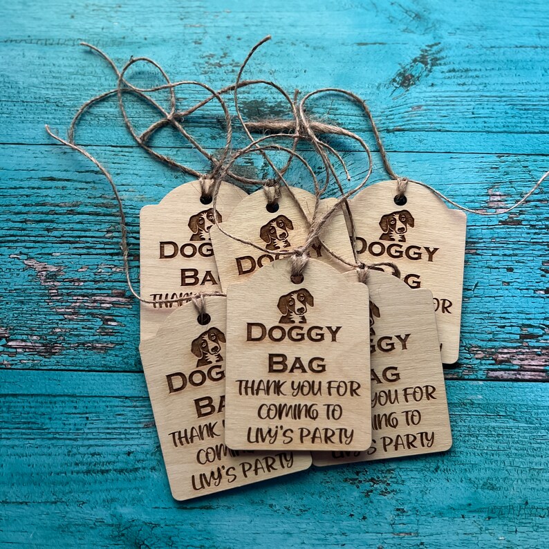 Peut inclure: Plusieurs &eacute;tiquettes cadeaux en bois avec une illustration de chien et les mots "DOGGY BAG" et "THANK YOU FOR COMING TO LIVY'S PARTY" grav&eacute;s. Chaque &eacute;tiquette est attach&eacute;e avec de la ficelle.