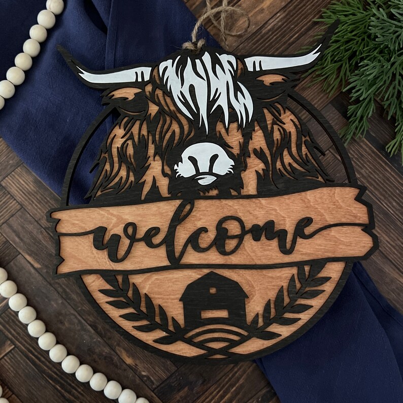 May include: Wooden welcome sign with a black silhouette of a highland cow with white markings and the word "welcome" in a decorative font.