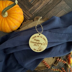 May include: A round wooden ornament with the text "Friends are the family Happy Friendsgiving 2023 That you pick" and a drawing of a pumpkin and fall leaves.