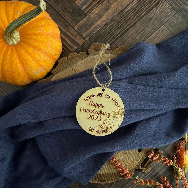 May include: A round wooden ornament with the text "Friends are the family Happy Friendsgiving 2023 That you pick" and a drawing of a pumpkin and fall leaves.