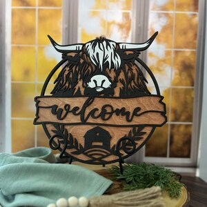 May include: A wooden welcome sign with a black silhouette of a Highland cow's head and the word "welcome" in a decorative font. The sign is on a black metal stand and has a brown wood grain background.