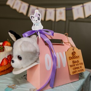 Peut inclure: Une bo&icirc;te rose pour cadeaux de f&ecirc;te avec une t&ecirc;te de chien en peluche. L'&eacute;tiquette en bois dit "Doggy Bag Thank You for Coming to Livy's Party." Un g&acirc;teau aux fraises et une banni&egrave;re sont en arri&egrave;re-plan.