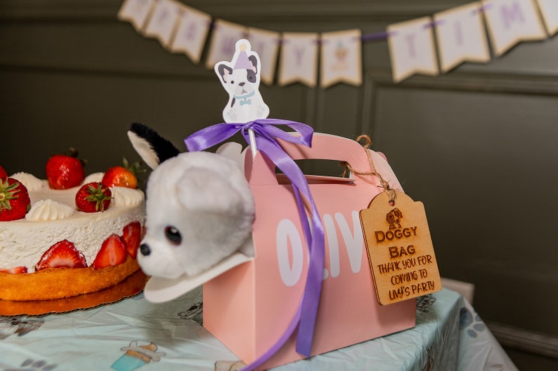 Peut inclure: Une bo&icirc;te rose pour cadeaux de f&ecirc;te avec une t&ecirc;te de chien en peluche. L'&eacute;tiquette en bois dit "Doggy Bag Thank You for Coming to Livy's Party." Un g&acirc;teau aux fraises et une banni&egrave;re sont en arri&egrave;re-plan.