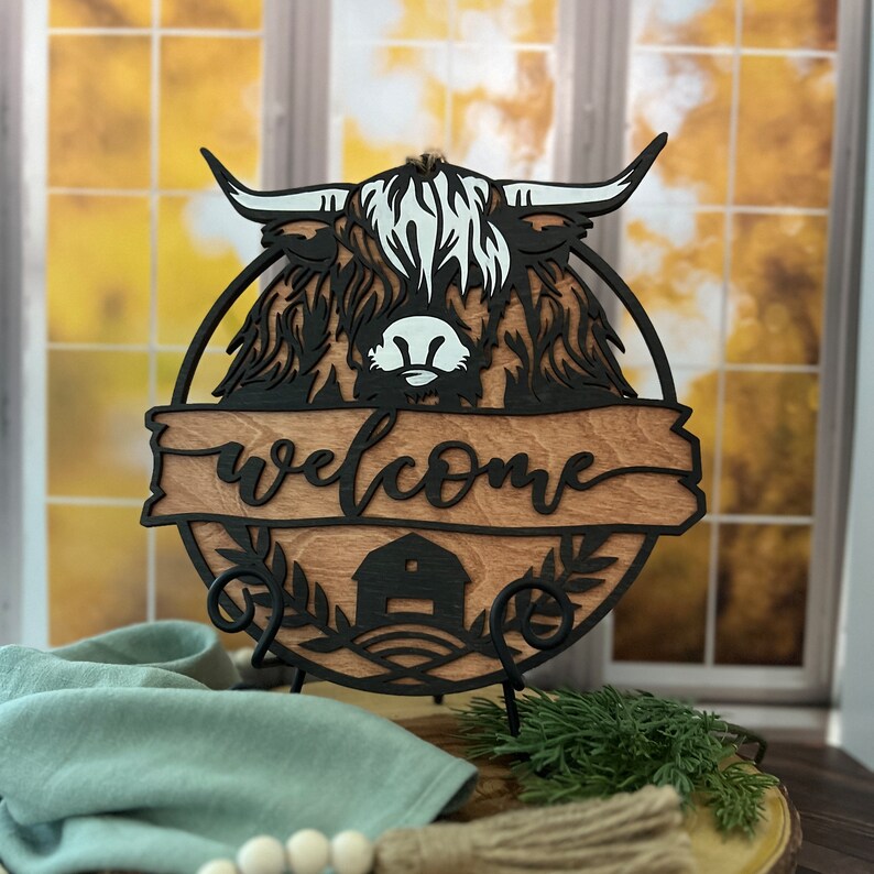 May include: A wooden welcome sign with a black silhouette of a Highland cow's head and the word "welcome" in a decorative font. The sign is on a black metal stand and has a barn and foliage design.